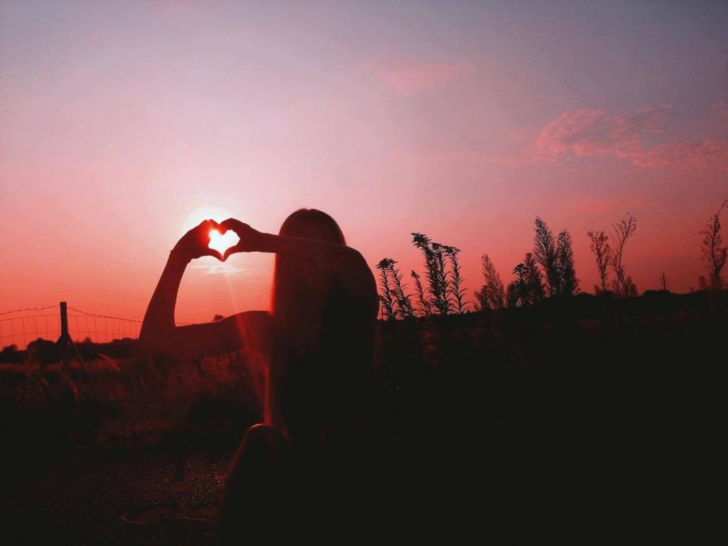 Woman making her hands into a heart against the setting sun.
